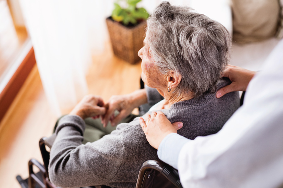 An older woman sitting in a wheelchair looking out a window. Behind her, someone is resting their hands on her shoulders