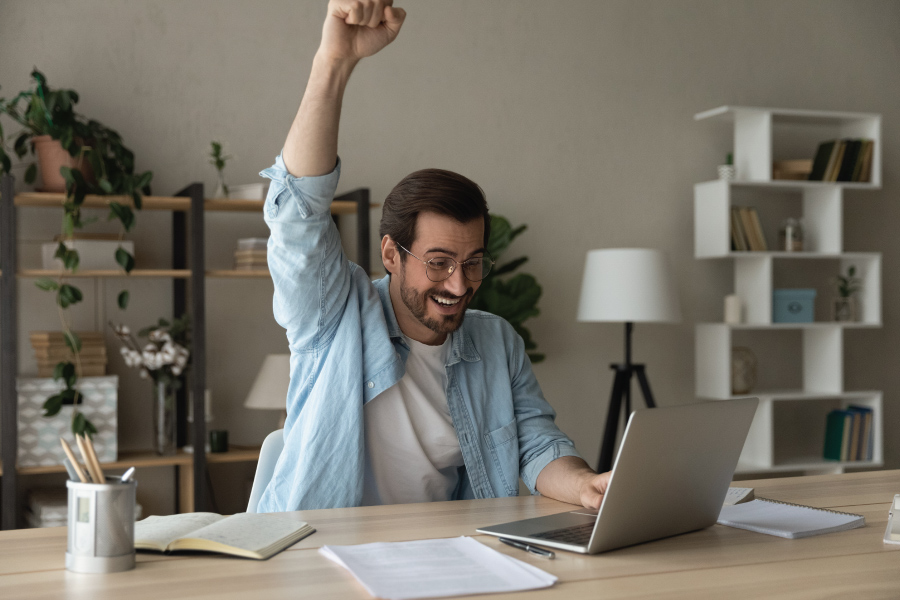 A man sitting at a desk with an open laptop is holding a fist in the air
