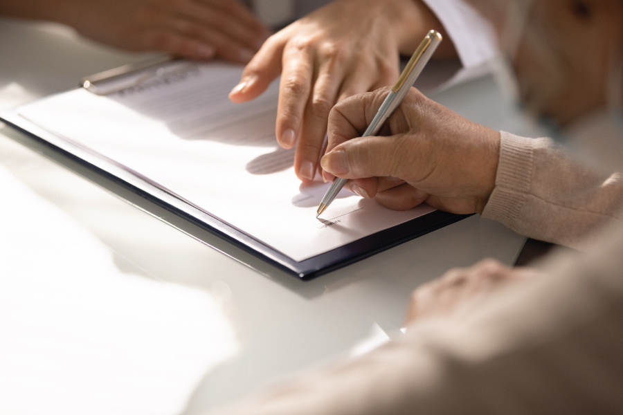 A hand signing a piece of paper. Another person's hand is pointing to the paper