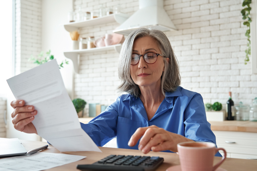 A woman using a calculator and holding a piece of paper sitting in a kitchen